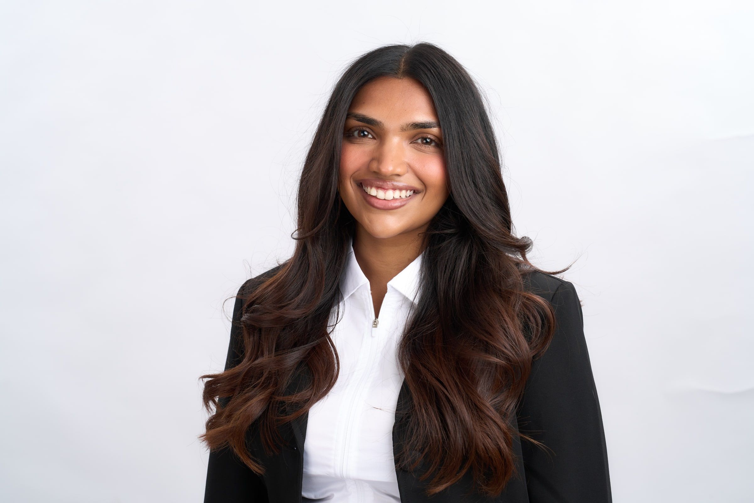 Delcy Pandya, smiling, in a black blazer and white shirt against a pale studio backdrop